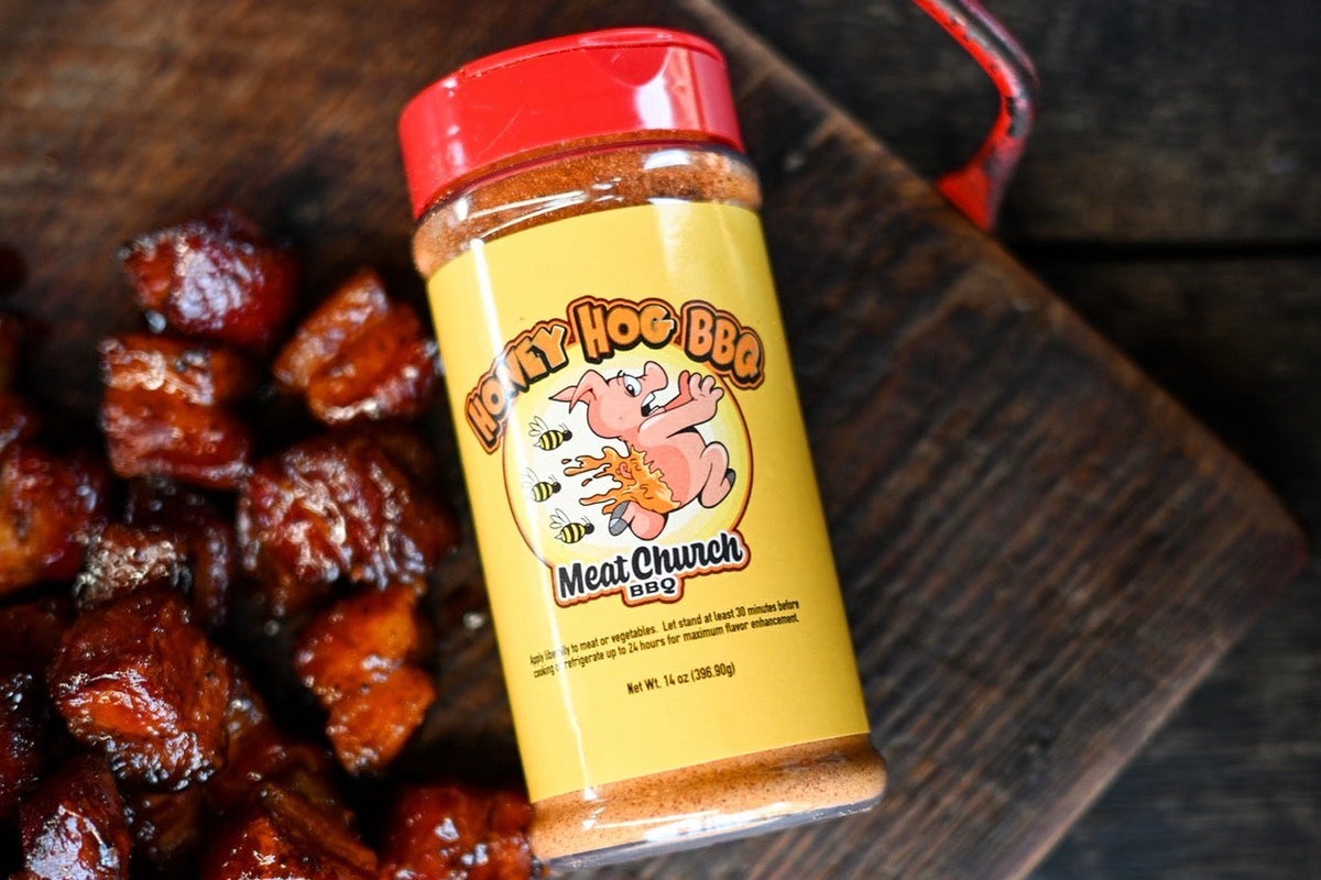 Meat Church Honey Hog BBQ rub shaker next to burnt ends on a dark wood cutting board