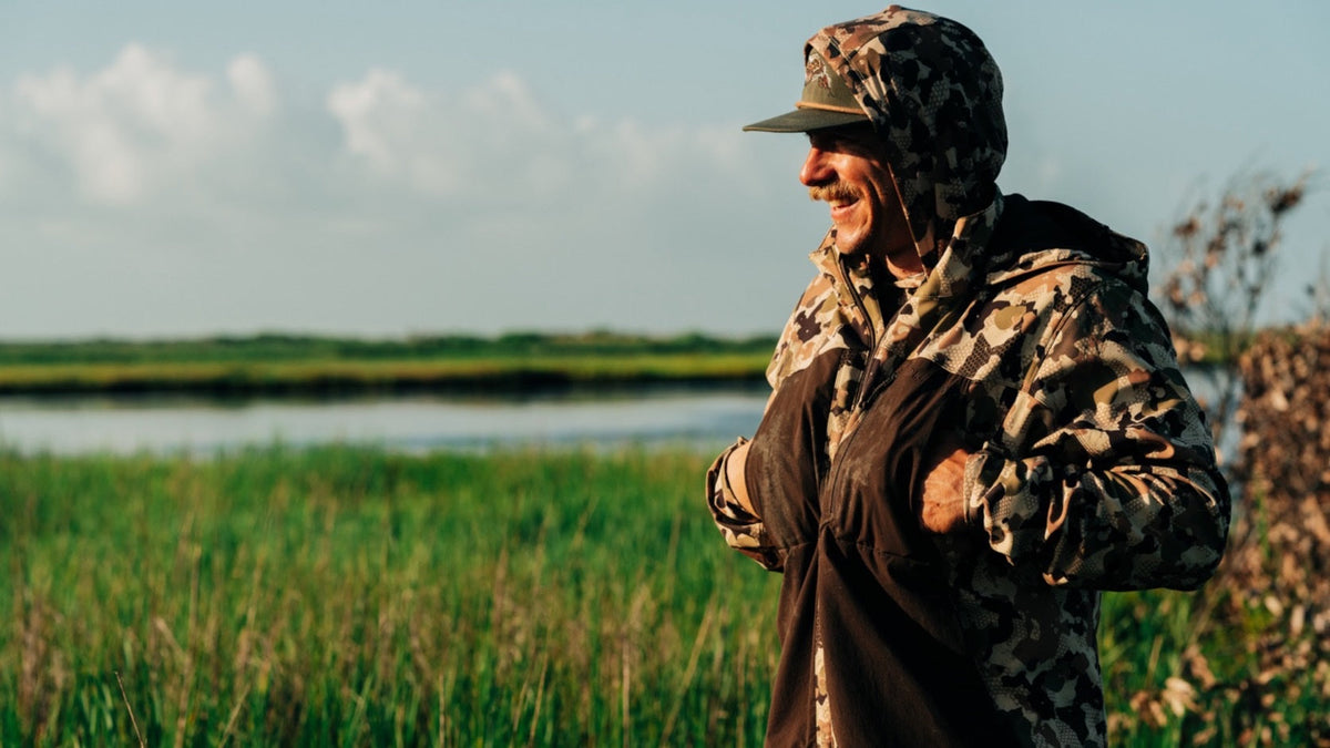 Duck Camp F26 — hunter in a camo hooded jacket standing in marsh grass
