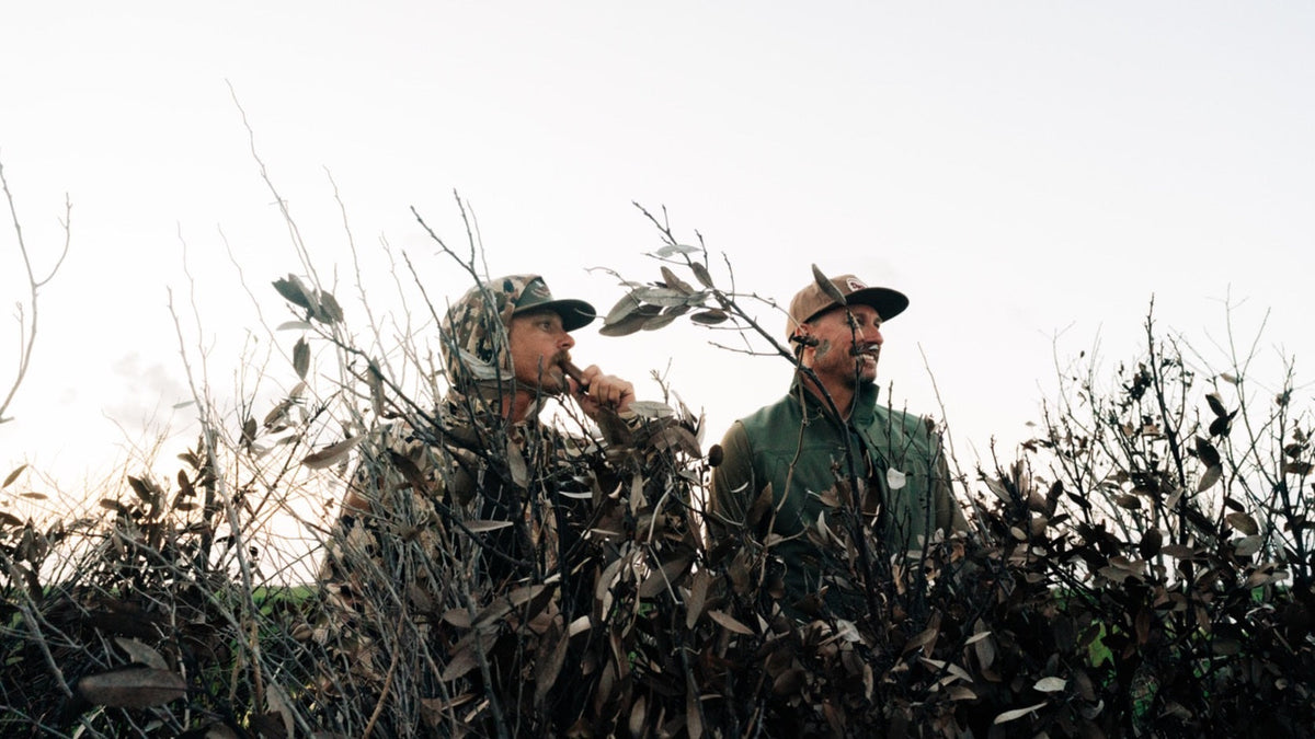 Duck Camp F26 — two hunters calling from a brush blind at first light