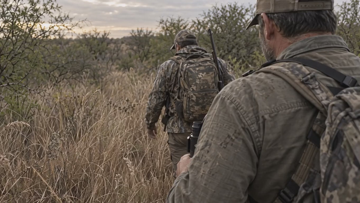 Load video: Two hunters in worn camouflage move through dry Texas grass, filmed from behind.